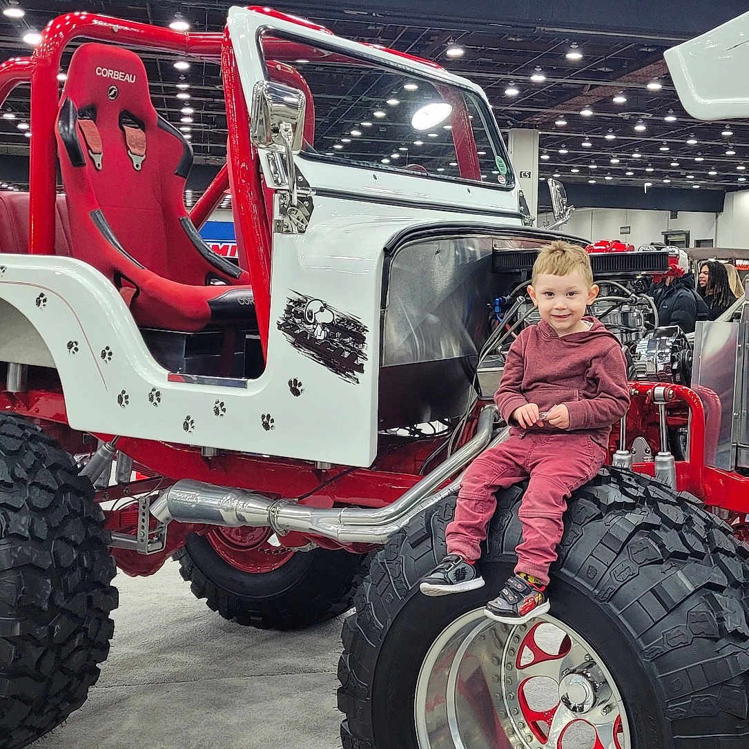 Ian is registered to the contest to win money with this photo: car_show, child, clothing, custom_car, engine, exhibition, floor, headlights, hood_open, indoor, large_tire, metal, offroad_vehicle, people, person, red_and_white, seat, smiling, sneakers, tire_tread