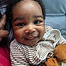 baby, infant, child, smiling, portrait, close_up, curly_hair, brown_skin, striped_clothing, onesie, stuffed_toy, orange_toy, cuddling, adult_hand, indoor, seated, happy, face, baby_photo, soft_lighting