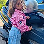 child, curly_hair, pink_jacket, blue_jeans, motorcycle, helmet, outdoor, vehicle, leaning, portrait, casual, expression, young, daylight, street, person, clothing, transportation, jeans, jacket
