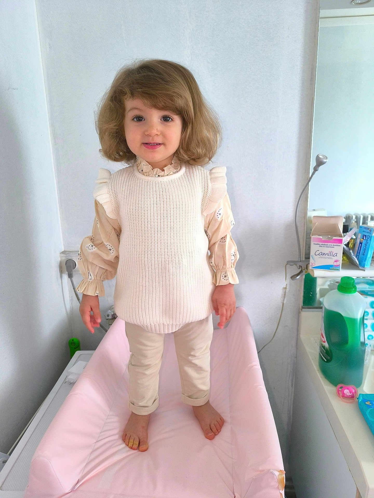 Kamylia a rejoint le concours — aidez-le/la à gagner de superbes lots ! child, toddler, standing, indoor, clothing, sweater, pants, barefoot, changing_table, mirror, bathroom, household_items, plastic_bottle, pacifier, face, hair, smile, person, wall, floor