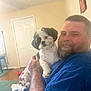 Buddy is registered to the contest to win money with this photo: man, dog, indoor, smile, beard, tattoo, blue_shirt, living_room, carpet, door, table, wall_art, furniture, pet, holding, happy, portrait, cozy, cute, fur