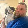 man, dog, indoor, beard, blue_shirt, holding, pet, calm, portrait, furniture, wall, door, tattoo, short_hair, closeup, light, home, person, animal, companion