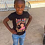 child, girl, smiling, standing, hands_on_hips, black_tshirt, graphic_tee, jeans, ripped_jeans, sandals, outdoor, bench, concrete_floor, sunny, building, window, hair_braids, casual_clothing, portrait, happy