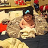 baby, infant, beanie, hat, pillow, bed, blanket, dinosaur_pattern, room, caps, valentines_day_card, stuffed_animal, toys, clothing, face, child, indoor, person, cozy, home