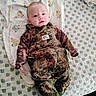 baby, child, camouflage, clothing, overalls, bed, blanket, pattern, floral, animal_print, person, infant, cute, face, eyes, expression, soft, lying_down, indoors, cozy