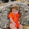 Cylis is registered to the contest to win money with this photo: baby, infant, child, hat, onesie, orange_clothing, pacifier, dinosaur_pattern, pillow, camo_hat, indoor, cozy, blanket, sitting, cute, curious, person, face, footwear, sock