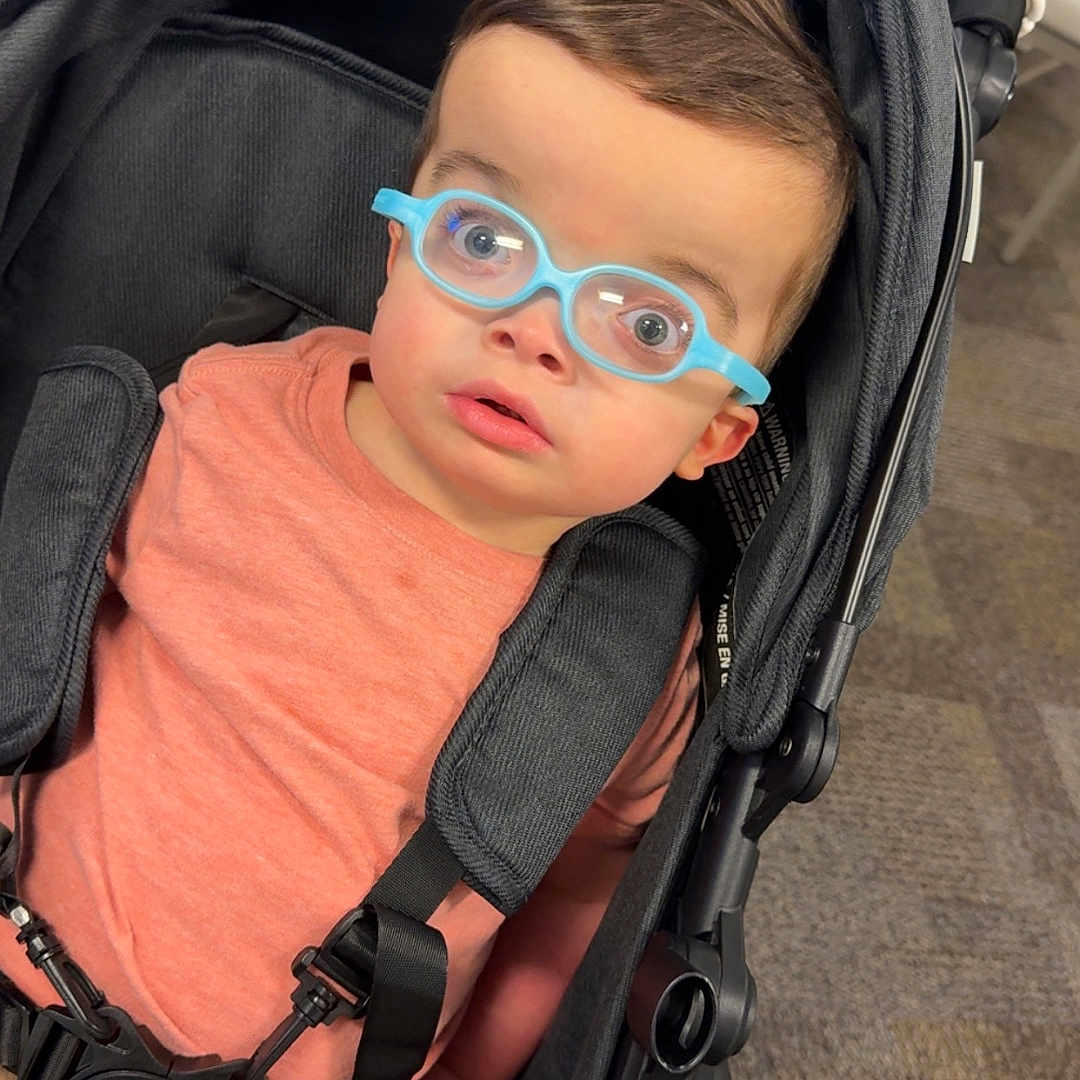Harvee joined the competition — help win amazing prizes! baby, child, toddler, stroller, glasses, blue_glasses, portrait, indoors, seated, sitting, looking_at_camera, wide_eyed, brown_hair, coral_shirt, pants, harness, carpet, playful, close_up, photography