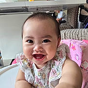 Janae is registered to the contest to win money with this photo: baby, smiling, child, high_chair, pink, floral_clothing, indoor, person, seated, table, plastic_bottle, ceiling_fan, face, happy, cute, infant, hand, furniture, home, bright