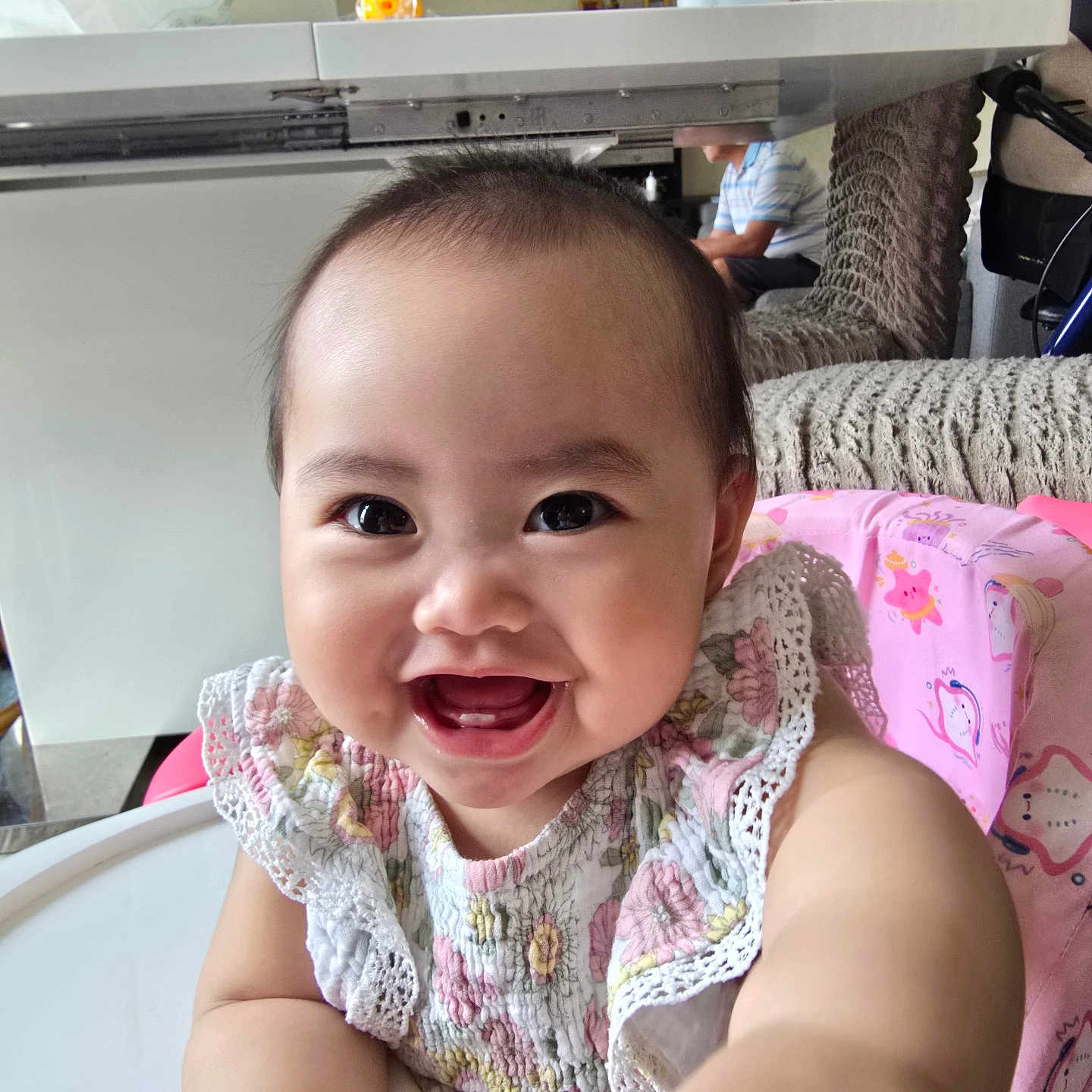 Janae is registered to the contest to win money with this photo: baby, smiling, child, high_chair, pink, floral_clothing, indoor, person, seated, table, plastic_bottle, ceiling_fan, face, happy, cute, infant, hand, furniture, home, bright