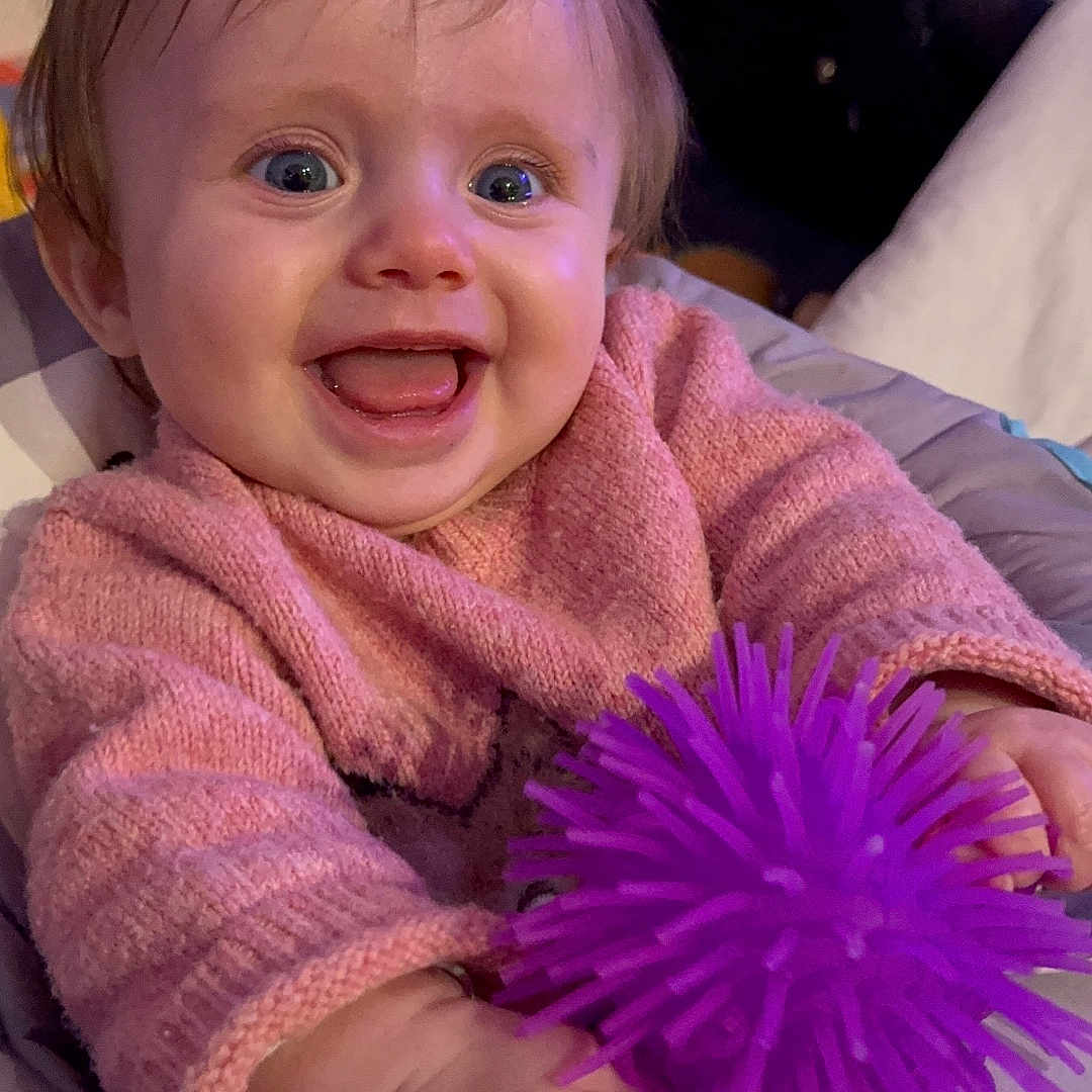 Amandine participe au concours pour gagner de l'argent avec cette photo : baby, bodypart, clothing, dahlia, daisy, face, finger, flower, hand, happy, head, knitwear, laughing, person, photography, plant, portrait, purple, smile, sweater
