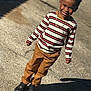 Lester V joined the competition — help win amazing prizes! child, boy, smiling, striped_shirt, brown_pants, black_shoes, outdoor, sunny, shadow, pavement, happy, casual_clothing, standing, portrait, daylight, fashion, accessory, necklace, curly_hair, person