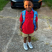 Lester V joined the competition — help win amazing prizes! child, boy, smiling, backpack, red_shirt, shorts, sneakers, standing, outdoor, driveway, car, pavement, grass, fence, daylight, portrait, person, happy, casual_clothing, school