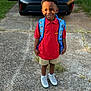 child, boy, smiling, backpack, red_shirt, shorts, sneakers, standing, outdoor, driveway, car, pavement, grass, fence, daylight, portrait, person, happy, casual_clothing, school