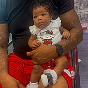 Emora is registered to the contest to win money with this photo: baby, child, man, tattoo, black_shirt, red_shorts, hello_kitty, shoes, socks, indoor, store, shelf, pink_items, person, holding, cute, curious, lap, seated, casual