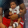 baby, child, man, tattoo, black_shirt, red_shorts, hello_kitty, shoes, socks, indoor, store, shelf, pink_items, person, holding, cute, curious, lap, seated, casual