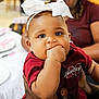 Kru is registered to the contest to win money with this photo: baby, blurred_background, child, closeup, clothing, cute, expression, eyes, face, hand_in_mouth, headband, holding, indoor, infant, maroon_shirt, person, portrait, skin, table, white_bow