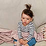 toddler, child, striped_shirt, blue_jeans, socks, blanket, couch, indoors, hair_tied, sitting, cross_legged, young_child, cute, portrait, casual_clothing, relaxed, comfort, home, person, face