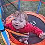 toddler, child, trampoline, netting, red_jacket, grass, outdoor, play, happy, smiling, safety_net, blue_pole, orange_padding, young_child, fun, clothing, footwear, person, daylight, recreation