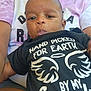 baby, child, person, infant, shirt, text, black_clothing, tie_dye, adult, holding, face, expression, outdoor, skin, head, eyes, mouth, hand, sitting, closeup