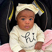 Leilani is registered to the contest to win money with this photo: baby, infant, child, car_seat, headband, bow, blanket, clothing, cute, face, portrait, newborn, sitting, indoor, expression, hands, pink_blanket, black_background, warning_label, apparel