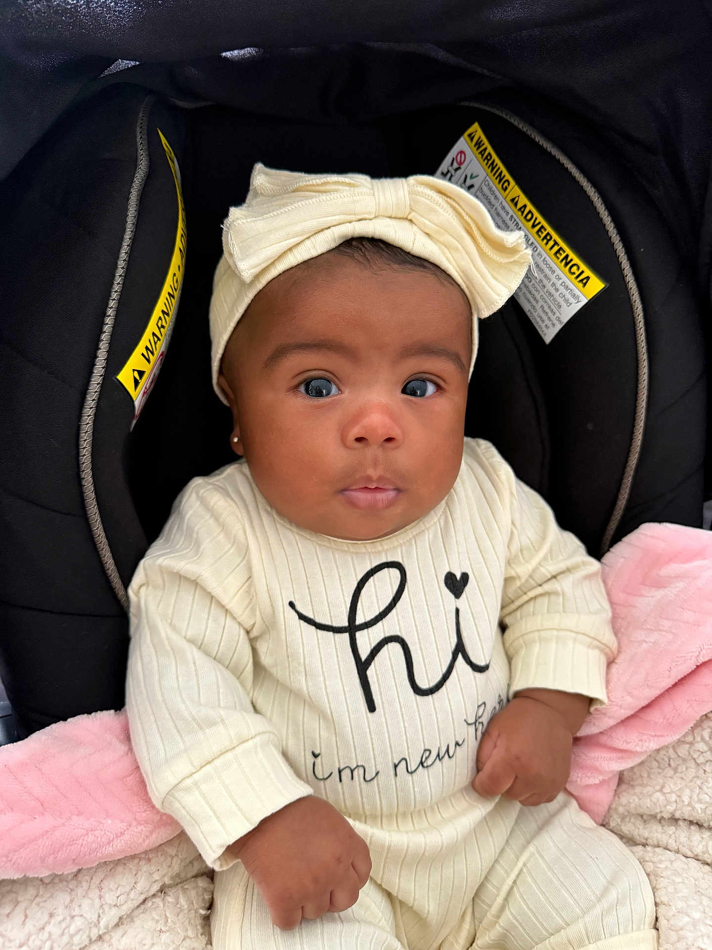 Leilani is registered to the contest to win money with this photo: baby, infant, child, car_seat, headband, bow, blanket, clothing, cute, face, portrait, newborn, sitting, indoor, expression, hands, pink_blanket, black_background, warning_label, apparel