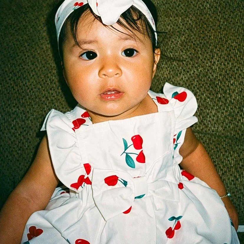 Sofia is registered to the contest to win money with this photo: toddler, child, dress, headband, cherry_pattern, white_dress, sitting, couch, green_couch, indoor, portrait, cute, baby, fashion, clothing, person, face, expression, young_child, headwear