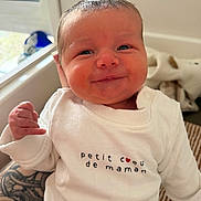 Louka a rejoint le concours — aidez-le/la à gagner de superbes lots ! baby, newborn, smile, white_clothing, onesie, tattoo, hand, face, infant, person, window, natural_light, cute, indoors, portrait, skin, expression, human, clothing, happy