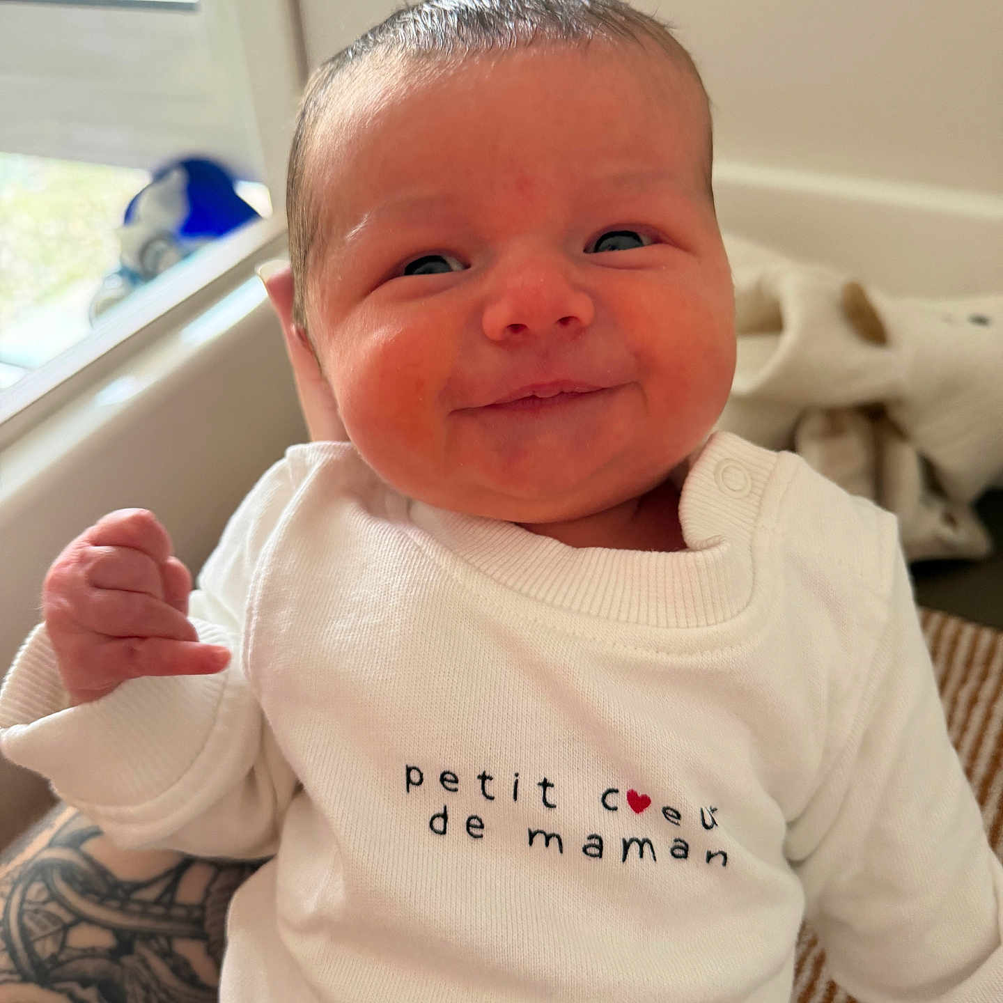 Louka a rejoint le concours — aidez-le/la à gagner de superbes lots ! baby, newborn, smile, white_clothing, onesie, tattoo, hand, face, infant, person, window, natural_light, cute, indoors, portrait, skin, expression, human, clothing, happy