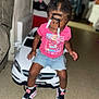 child, toddler, toy_car, indoor, braided_hair, beads, pink_shirt, denim_shorts, sneakers, floor, car, playing, cute, young_child, person, casual_clothing, fun, small_vehicle, looking_down, home