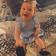 Rylee is registered to the contest to win money with this photo: baby, child, smiling, blue_dress, bow, bed, blanket, footwear, flip_flop, happy, indoor, person, plaid_pillow, patterned_bedding, cute, portrait, sitting, young_child, home, cozy