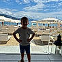 beach, boy, child, cloud, daylight, lounge_chair, outdoor, person, relaxation, sand, sandal, sea, shore, shorts, sky, standing, summer, tshirt, umbrella, vacation