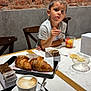 brick_wall, chair, child, coffee, croissant, cup, drink, food, fork, fruit_salad, indoor, marble_table, napkins, person, plate, restaurant, snack, sugar_packets, table, tile_wall