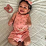 baby, child, smile, pink_clothing, headband, toy, blanket, footwear, indoor, cute, happy, infant, face, lying_down, patterned_clothing, accessory, cherry_pattern, playtime, soft_texture, shoes