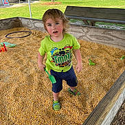 Waylon joined the competition — help win amazing prizes! child, toddler, corn_kernels, corn_pit, green_shirt, tmnt_shirt, shovel, toy_shoes, play_area, wooden_box, bench, grass, tent, outdoor, playground, messy_hair, curious_expression, face, smile, daytime