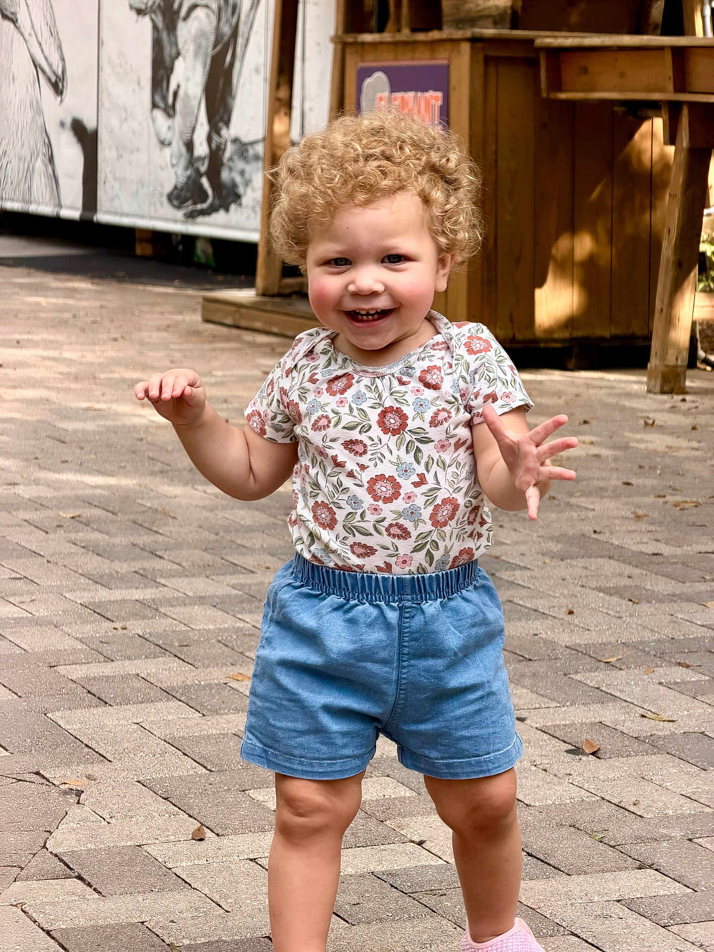 Isabella is registered to the contest to win money with this photo: toddler, child, curly_hair, smiling, outdoor, pavement, floral_shirt, blue_shorts, walking, happy, person, daylight, sunlight, cute, young_child, casual_clothing, shoes, summer, playful, nature