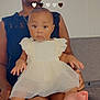 Isla is registered to the contest to win money with this photo: baby, child, adult, woman, sitting, dress, white_dress, lace, indoor, heart_emojis, expression, face, legs, hands, foot, necklace, earrings, chair, couch, skin