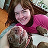 Ellie May is registered to the contest to win money with this photo: woman, dog, smiling, happy, indoor, bed, cuddling, pet, portrait, casual, brown_hair, pink_shirt, wooden_furniture, photo_frame, playful, teeth, closeup, relaxing, cozy, home