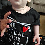 Christian joined the competition — help win amazing prizes! baby, infant, child, face, eyes, mouth, t_shirt, heart_symbol, hand, couch, living_room, blanket, carpet, gray_pants, sitting, portrait, curious_expression, close_up, skin, person