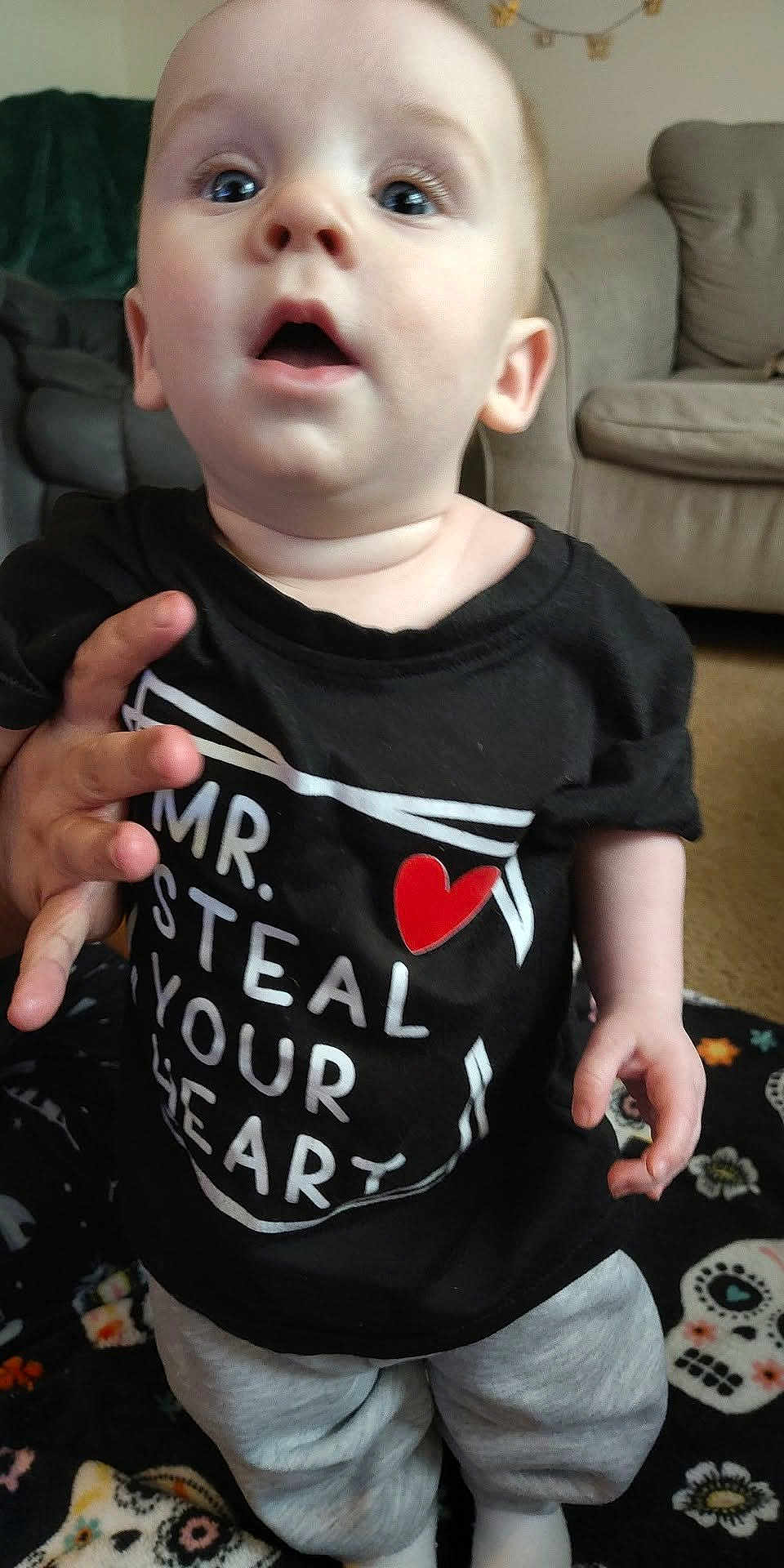 Christian joined the competition — help win amazing prizes! baby, infant, child, face, eyes, mouth, t_shirt, heart_symbol, hand, couch, living_room, blanket, carpet, gray_pants, sitting, portrait, curious_expression, close_up, skin, person