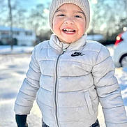 Sebastian is registered to the contest to win money with this photo: child, smiling, winter_clothing, puffer_jacket, knit_hat, pom_pom, outdoor, snow, gloves, camo_pants, happy, cold_weather, face, person, daylight, background_blur, nature, vehicle, casual, portrait