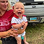 baby, woman, smile, happy, outdoor, grass, truck, ford, license_plate, summer, casual_clothing, holding, child, red_shirt, shorts, pacifier, blue_eyes, light_hair, cheerful, daylight