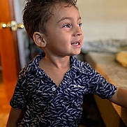 Carmelo is registered to the contest to win money with this photo: child, boy, smile, curly_hair, shirt, patterned_clothing, indoor, door, table, furniture, face, portrait, happy, young, person, skin, eye, mouth, hair, background_blur