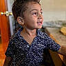 child, boy, smile, curly_hair, shirt, patterned_clothing, indoor, door, table, furniture, face, portrait, happy, young, person, skin, eye, mouth, hair, background_blur