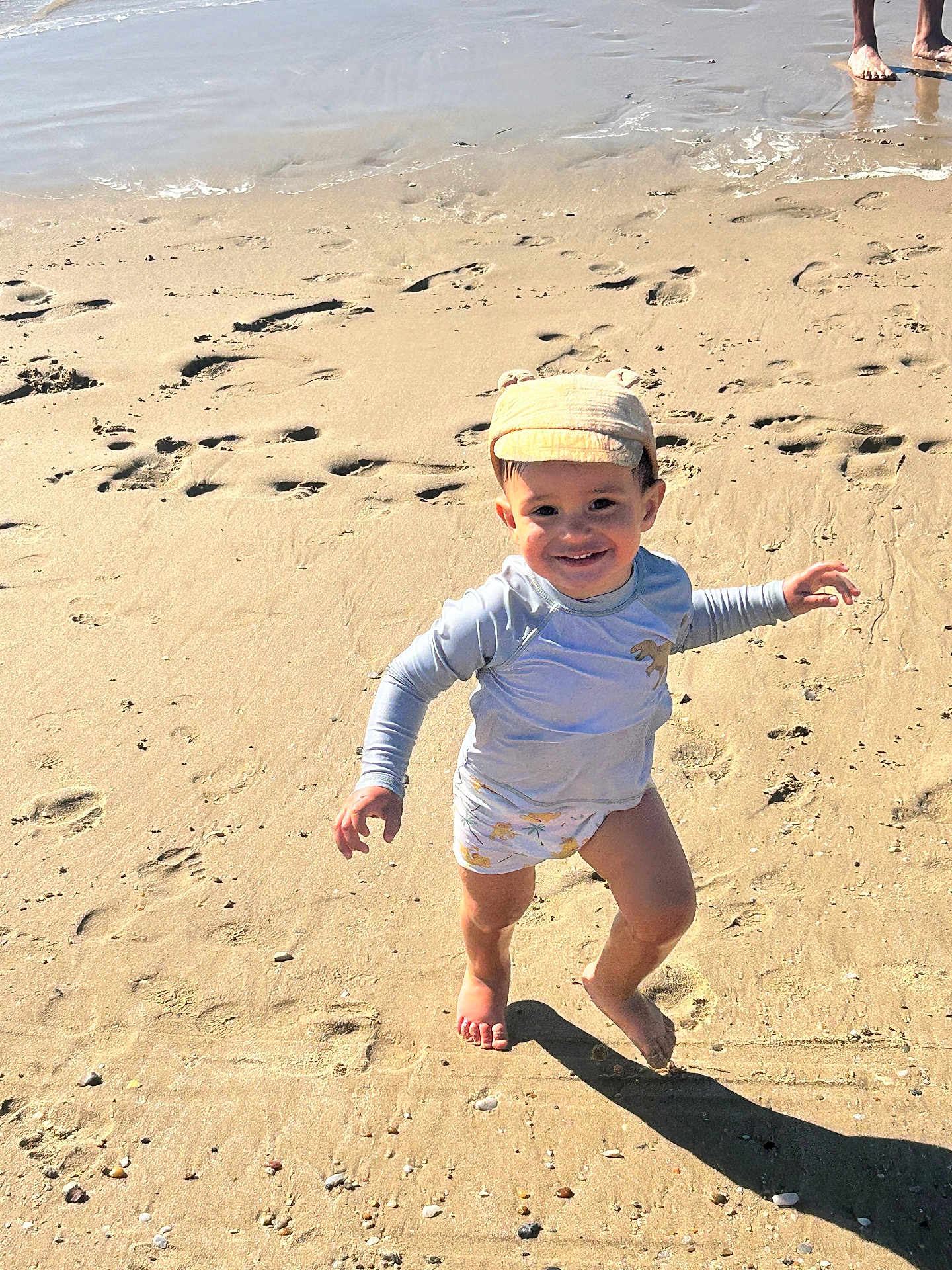 Gaspard a rejoint le concours — aidez-le/la à gagner de superbes lots ! child, toddler, beach, sand, barefoot, hat, running, smiling, water, footprints, sunlight, shadow, shore, playful, summer, clothing, person, outdoor, happy, nature