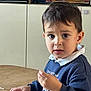 child, boy, indoor, table, sweater, face, hand, snack, curious, expression, wooden_table, hair, clothing, portrait, young, person, casual, real_people, sitting, looking