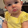 baby, child, yellow_dress, headband, bow, indoor, portrait, cute, infant, expression, seated, furniture, soft_lighting, person, baby_girl, clothing, face, hands, closeup, curious