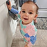 toddler, child, smiling, barefoot, colorful_clothing, kitchen, cabinet, floor, patterned_rug, person, indoors, curly_hair, happy, young_child, holding, knob, standing, cute, face, eyes