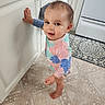 toddler, child, barefoot, standing, kitchen, floor, cabinet, hand, looking_up, curious, floral_outfit, patterned_floor, appliance, indoors, young_child, person, face, head, skin, expression