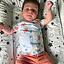 baby, child, infant, lying_down, shirt, shorts, cushion, star_pattern, raised_arm, face, person, indoors, cute, smiling, young_child, clothing, bed, resting, toddler, happy