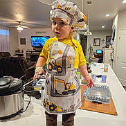 Nakoa joined the competition — help win amazing prizes! apron, barefoot, brown_pants, ceiling_lamp, chef_hat, child, countertop, curtains, fan, indoor, kitchen, living_room, person, plant, plastic_container, pressure_cooker, television, toddler, wooden_cutting_board, yellow_shirt